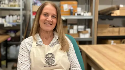 A woman in an apron, bearing the branding Nantwich Candle Co. She is sat on a chair by a table, and is smiling at the camera
