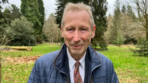 Roger Deeks wearing a patterned tie and white collared shirt underneath a dark blue raincoat. He has short dark blonde hair and is smiling at the camera. He is standing in the middle of a large green space dotted with mature trees. 