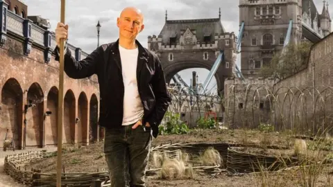 Landscape gardener and horticulturalist Nigel Dunnett wearing black jacket and trousers and white t-shirt, holding a stick with the Tower of London behind. Dunnett designed the wildflower project for Superbloom in the Tower of London moat, (date).