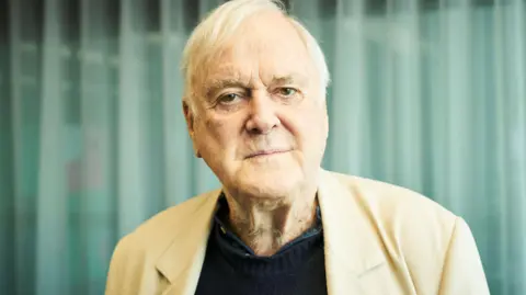 BBC / Jonathan Birch Comedian John Cleese is pictured wearing a beige suit jacket and a black woollen jumper. He is standing in front of some net curtains. He has brown eyes and white hair.