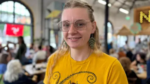 Celia Bales has long blonde hair, half tied up. She is smiling, wearing glasses and a yellow t-shirt. She is standing in a busy cafe with high ceilings.