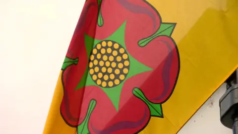 An image of a Lancashire flag flying. The flag is yellow with the Lancashire Red rose on it.