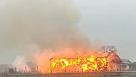 A picture taken from the A1 of the fire. The barn is engulfed in orange flames. Only the framework of the building remains standing. A large plume of smoke trails into the air.