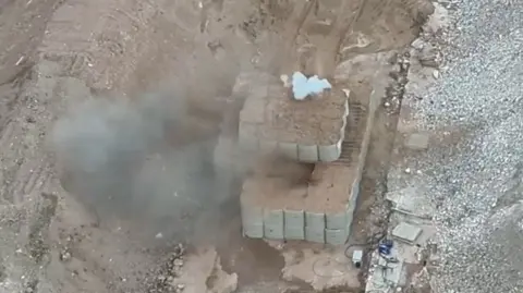 Smoke rises through dozens of giant sand bags as a bomb is detonated