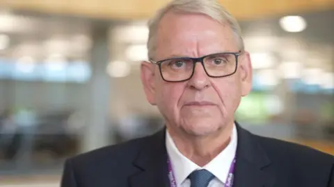 Zoie O'Brien/BBC A head and shoulders shot of a man with white hair and black-framed glasses, wearing a dark suit over a white shirt with a blue tie. He is standing in a room.