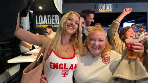 Two young women in England tops smile as they cheer for joy at Ashton Gate Stadium. Other people can be seen celebrating behind them.