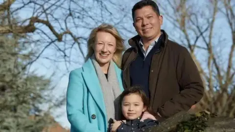 Surrey Police A woman with blonde hair stood to the left of a man with black hair. they are stood behind a young girl with brown hair. They are stood outside in front of brown trees. They are all smiling and facing the camera