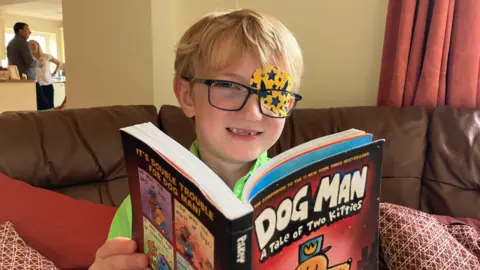 Harry is sitting on a couch reading a book called Dog Man. He is smiling at the camera wearing glasses and a patch over one eye with rockets and stars on it. 