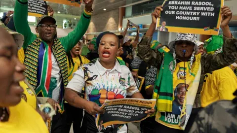 AFP/Getty Images Supporters of South Africa expelled ambassador to the US Ebrahim Rasool, many dressed in the yellow, green and black colours of the ANC, wait to greet him at the Cape Town International Airport on 23 March 2025. Some hold posters reading: 'Ebrahim Rasool, you have served our country with honour!!'