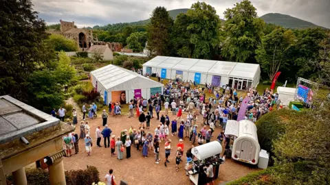 Lloyd Smith Photography An aerial view of the Borders Book Festival with food trucks, marquees and people milling about. It is surrounded by trees with Melrose Abbey in the background and hills in the distance