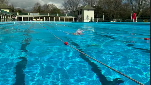 Peterborough lido, the pool is separated into lanes and there is one swimmer. In the distance the sky is overcast and grey.