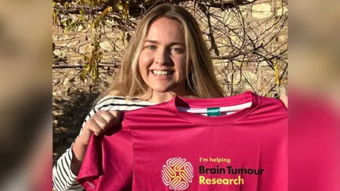 Megan Williams Megan smiling whilst holding up a pink t-shirt saying 'Brain Tumour Research'