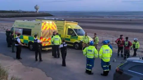 More than a dozen emergency service workers in high-visibility jackets and police uniforms wait near two ambulances. They are next to the sands of Crosby beach around sunset with the sea in the background.