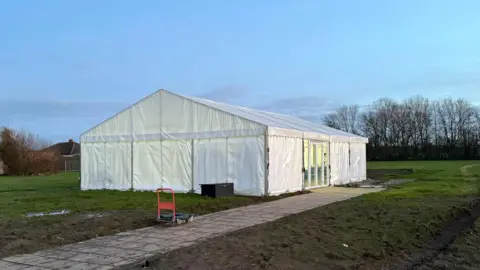 BBC A white gazebo-style marquee in the middle of a muddy grass field. The marquee has glass French doors on its right hand side. There is a light-coloured paved path leading up to the French doors. In the background, a blue sky and leafless trees. 