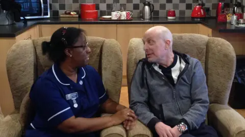 A nurse speaks to man while both are seated on armchairs.