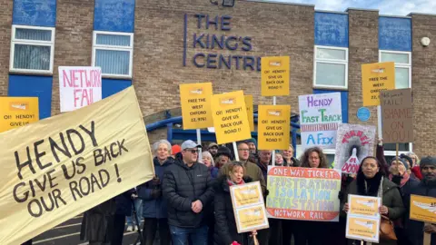 A group of protesters in front of The King's Centre in Oxford. They have placards and yellow banner reads "Hendy give us back our road!"