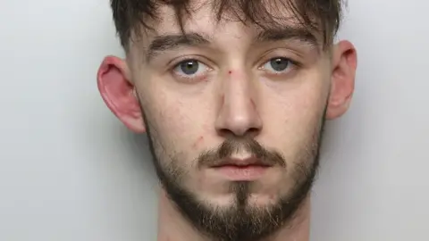 In a police custody photo, Jacob Power looks blankly at the camera. He has dark brown hair, a beard and prominent ears.