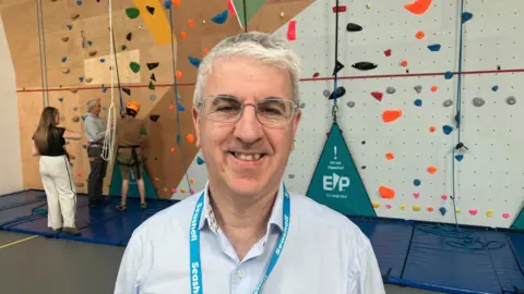 Photograph of Brandon Leigh who is the Chief Executive of the Seashell Trust in Stockport. He is pictured in front of a climbing wall at the college. 