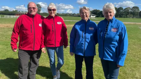 Bargain Hunt contestants. Alan and Shirley made up the red team and Carol and Cathy made up the blue team. All four of them are standing in a line wearing their team fleeces. They are looking at the camera and smiling. 