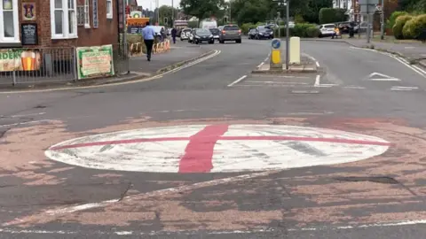 A red cross painting on a white mini roundabout, at a road junction with a scuffed red tarmac surface and black tyre marks on the white of the roundabout. A pub can also be seen, with pedestrians and cars in the background.