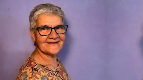 SUPPLIED A woman with short grey hair, black glasses and a blouse with autumn colours and flowery patterns stands in front of a purple wall. This is a head and shoulders photograph of the woman.