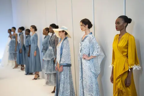 IHSAAN HAFFEJEE / GETTY IMAGES Women line up with their back against the wall of the catwalk. Most are dressed in shades of blue, while the woman at the front wears a mustard-yellow dress with elaborate white detailing at the cuffs.