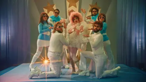 Eilís Doherty/AWAL A group of men and women in 70s attire surrounding a woman in a white top with a pink hood. They look to be standing on a blue stage.