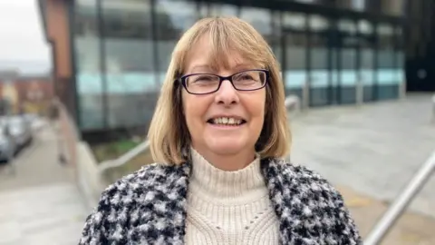 Laura Coffey/BBC Wendy Randall with medium-length brown hair and glasses, wearing a black and white cardigan outside a modern office building