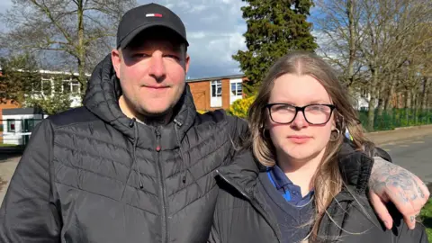 Shaun Whitmore/BBC Leon is wearing a black hooded coat and a black cap. He has his arm round Letizia, who is wearing a black coat over a V-neck jumper and navy polo shirt. She has long, light brown hair and is wearing glasses.