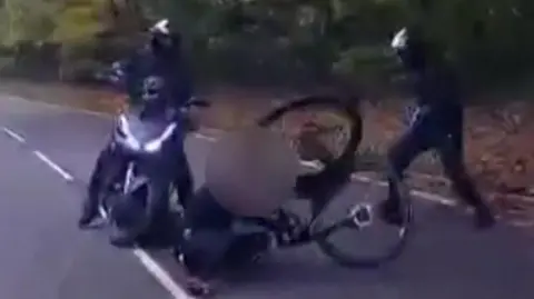 A gang stand over a man on the ground who has been knocked off his bike on a country road.