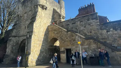 BBC/ Emily Johnson The Bootham Bar section of York's city walls, which includes a tower, two arches and a stone staircase. Tourists gather in front of it as a meeting point.