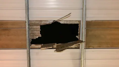 Image of a hole, taken from below, in a ceiling between two metal posts - showing damaged corrugated roofing hanging down into the opening and darkness above.