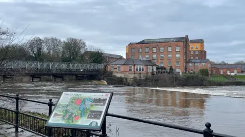 A swollen river with a bridge and mill in the background
