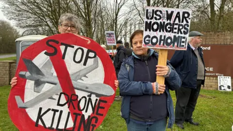Two women stand in front of other protesters near an RAF Fairford entrance sign. They are holding signs, one is round and in the format of a giant stop sign in red. It features a painted military drone and the words "Stop drone killing". The other sign simply states "US war mongers go home" in black capital letters.