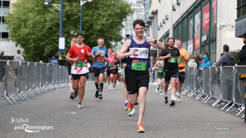 Dave Pullin/Marathon Photos Live A man with brown hair is running along a street with metal barriers on either side. He is wearing a purple vest with a large pink and purple "C" on it. it reads "Cancer Research UK". lower on the vest is a green running bib that says "Dave, 2049". He is smiling and he has his thumb up. There are people running behind him