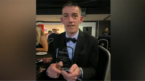 James Kelly Image of James, in suit and bow tie at an awards ceremony, holding a glass trophy. 