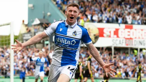 Getty Images Elliot Anderson smiles and holds his arms out as he runs away from goal after scoring for Bristol Rovers at the Memorial Stadium. Fans can be seen celebrating in the main stand behind him
