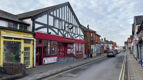 Johnny Right/BBC Liston High Street. Next to the cinema is a yellow colored pottery studio, with a burgundy colored front and glass windows with steps leading to the entrance. 