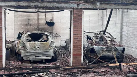 Two cars inside a garage that are squashed and destroyed by fire. The roof has fallen in and there are bricks and rubble on top of and around the cars