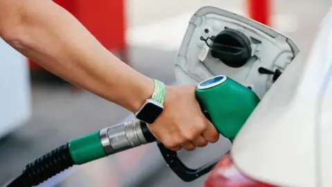 A close up stock image of an arm with a smartwatch with a light green strap putting a green unleaded petrol pump handle in to a car. The car is white.
