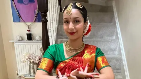 Saieesha Bailoor A woman with traditional Indian dance clothing, stood in front of some grey carpeted stairs inside.