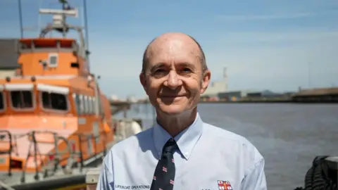 Shaun Whitmore/BBC A man wearing a blue shirt and a navy tie. He is smiling at the camera 