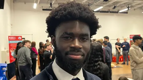 6th former at Holcombe Grammar School, Peniel Duah, stands in school hall whilst fellow pupils perform CPR on resuscitation dolls