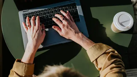 Catherine Falls Commercial/Getty Creative A woman sits at a laptop with her hands over the keyboard and a disposable cup and lid beside her.