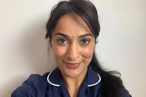 Harpreet Matharu A selfie taken by a nurse wearing blue scrubs. She looks at the camera and smiles, while standing in front of a white background.