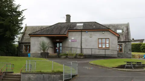 A grey school building with a small green play area in front, a tree beside it and a grey sky overhead.