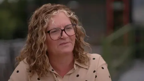 Catherine Marchant has shoulder length blonde wavy hair and black rimmed glassed. She wears a beige shirt with black polka dots. She looks deep in thought, away from the camera.