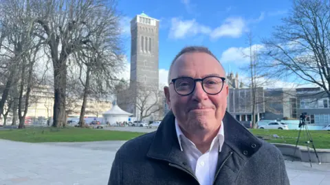 Plymouth City Council leader Tudor Evans, a middle-aged man with short hair, glasses, is standing outdoors with the tower of the Plymouth Guildhall in the background. He is wearing a dark-coloured coat over a white shirt.