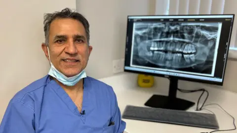 Shiv Pabary is pictured in his blue dental scrubs and wears a face mask around his neck. He is pictured next to image of an X-Ray of some teeth on his computer.