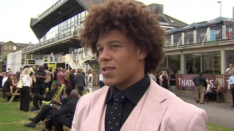 Buster Lawrence in a pink suit with a black shirt and polka dot tie standing outside the cricket club with many people behind him.